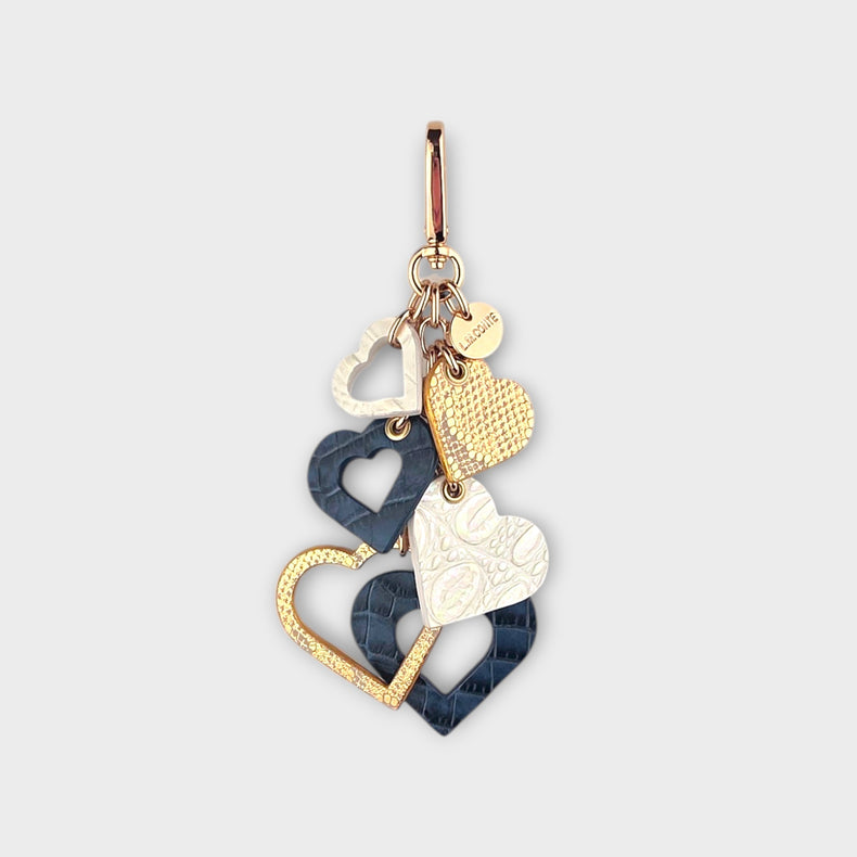 An LMCONTE multi-piece cluster of hearts handbag charm featuring textured gold, a warm pearlescent white, and denim blue crocodile-embossed leather hearts, suspended from a gold clip on a light gray background.