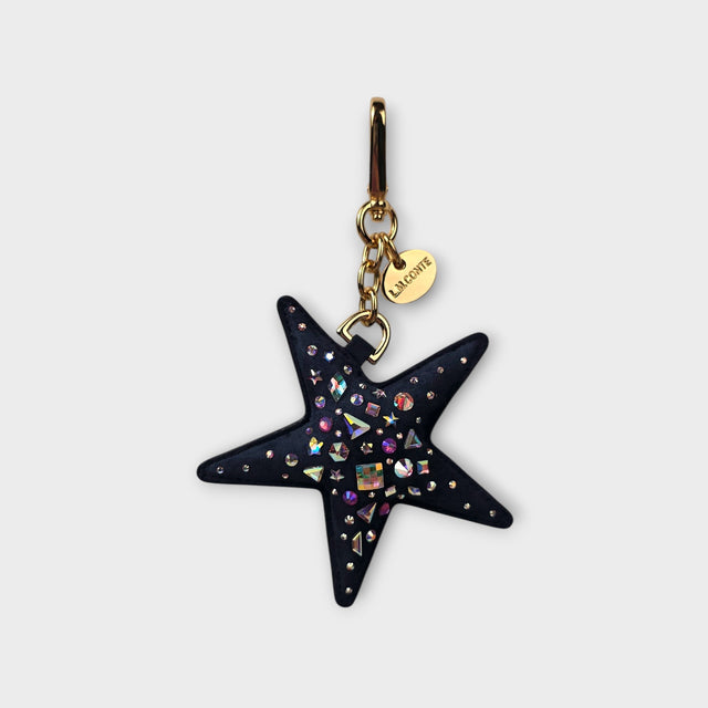 A dark navy blue suede celestial star charm embellished with iridescent crystal stones and gold-tone hardware, featuring a small gold tag engraved with 'LMCONTE'.