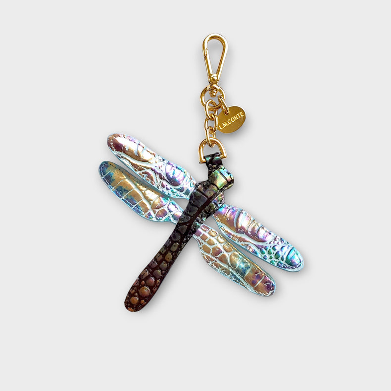 Dragonfly-shaped keychain with iridescent wings and gold accents photographed against a soft gray background.