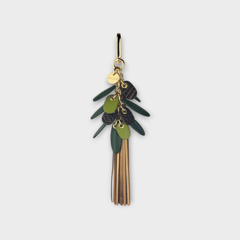 A handbag charm with green leaves and long fringe tassels in shades of gold against on a light gray background.