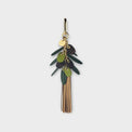 A handbag charm with green leaves and long fringe tassels in shades of gold against on a light gray background.