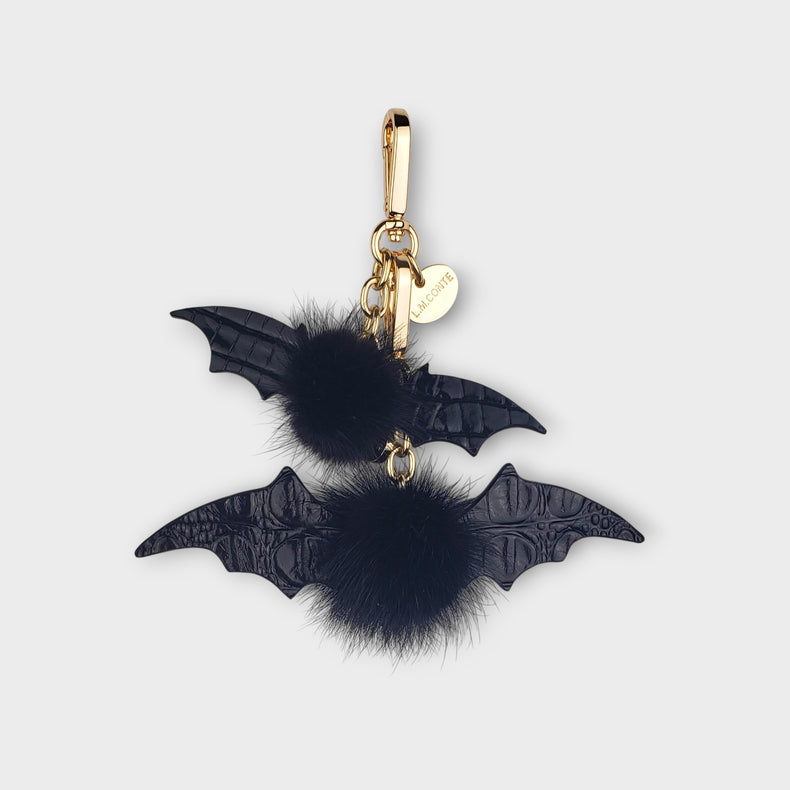 Two bat-shaped handbag charms with black mink fur pom-poms on a light gray background