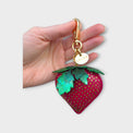 Strawberry-shaped purse charm held by a hand on a light gray background.