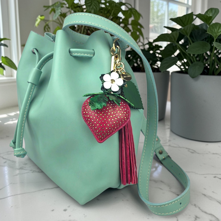 Mint green handbag with an L.M.CONTE strawberry bag charms on a marble surface with plants in the background.