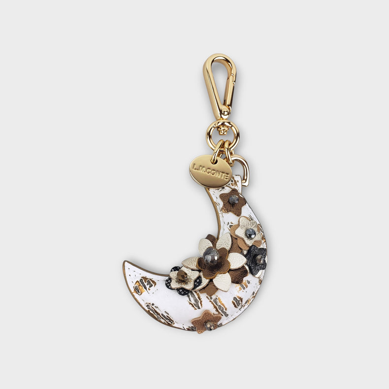 Decorative purse charm with 3D florals on a white heavily textured leather crescent moon shape with gold tone hardware photographed against a soft gray background.