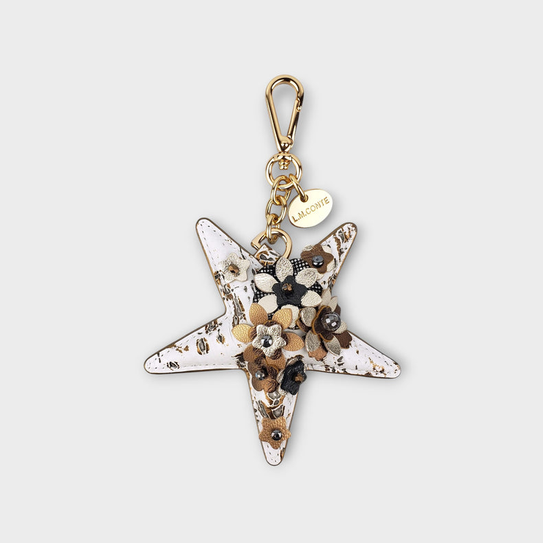 Decorative handbag charm with 3D florals and beaded embellishments on a white heavily textured leather star shape with gold tone hardware photographed against a soft gray background.